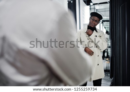 Stylish casual african american man at black beret and white trench raincoat at fitting room clothes store , looking at mirror.