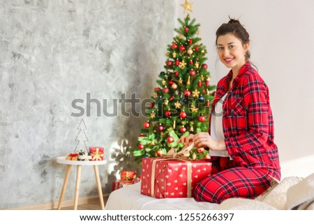Young beautiful brunette woman in plaid checkered pajamas in decorated bedroom interior with gift box and Chrictmas tree on background. People on Christmas morning concept. Close up, copy space.