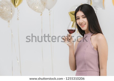 Attractive young asian woman in pink dress, holding her drink and happy at fun party, portrait on white background with colorful festive balloons.