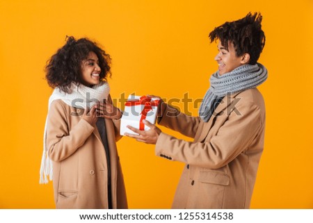 Happy african couple wearing winter clothes standing isolated over yellow background, giving presents