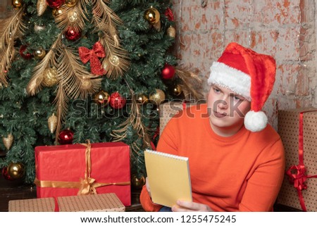 Handsome guy in christmas hat is writing a letter to Santa sitting under the tree surrounded by boxes of gifts. Christmas and gifts