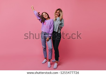 Slim joyful girls laughing during studio photoshoot. Indoor photo of emotional female friends isolated on rosy background.