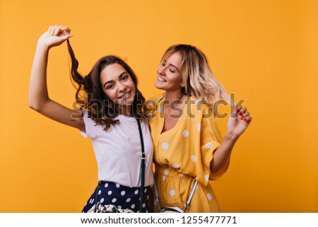 Adorable brunette girl touching her hair and laughing to camera. Blithesome caucasian sisters funny dancing on bright background.