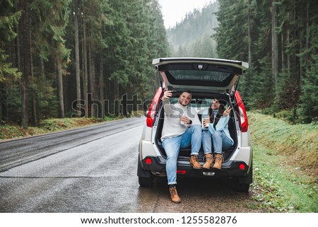 man and woman stop to drink warm up tea and taking selfie in car suv trunk. road trip concept