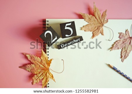 Christmas theme black painted wooden dice with the number 25 and December on white blank notebook paper with a pencil, dried and withered maple leaves over pink background. (top view, space for text)