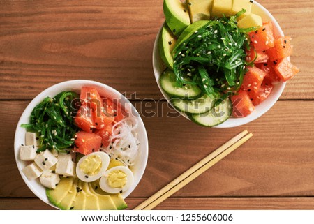 Raw Organic Poke Bowl with with rice, avocado, salmon, mango, cucumbers, chuka salad, quail eggs sweet onions with chopsticks two plates close-up on wooden background top view