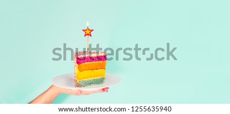 Female hand holding white plate with slice of Rainbow cake with birning candle in the shape of star isolated on blue background. Happy bithday, party concept. Wide banner. Selective focus. Copy space