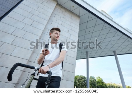 Young hipster style man posing with bicycle and listening music on the street, sport style picture, handsome guy with black backpack in the city at the background of a modern building