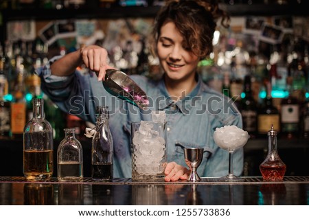 Smiling female bartender adding to the measuring glass cup an ice cubes with shovel on the bar counter on the blurred background