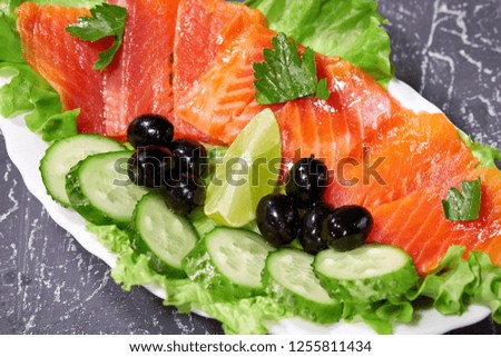 Sliced salmon pieces on a white plate with olives and lemon, on a grey background.