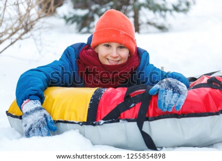 cheerful cute young boy in hat red scarf and blue jacket lays on tube on snow, has fun, smiles. Teenager on sledding in winter park. Active lifestyle, winter activity, outdoor winter games, snowballs