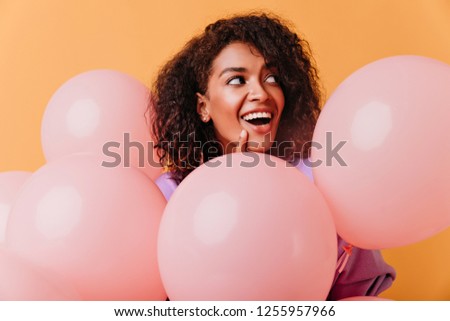 Amazing black female model with party balloons posing on orange background. Indoor photo of adorable brunette woman having fun during event.