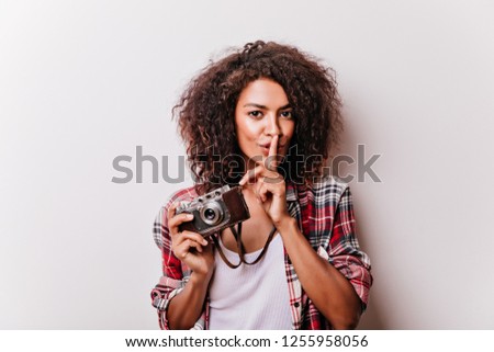 Good-humoured young lady with camera standing in studio with smile. Indoor photo of refined black girl taking pictures.