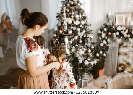 Сaring mother braids her little daughter's braid while second daughter decorates a New Year's tree in the light cozy room