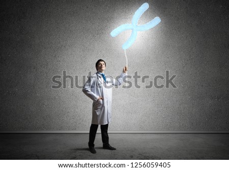 Conceptual image of successful and young doctor in white uniform interracting with DNA structure while standing inside empty roon with dark gray wall on background.