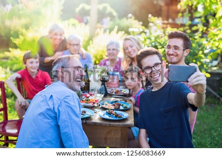 In the summer, a family of three generations gathered around a table in the garden sharing a meal. A teenager does a selfie with all the guests