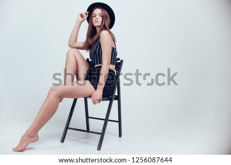 Attractive young model with beautiful make-up and hair-do in stylish black hat and black clothes posing with a chair on a white studio background. Long legs, brown hair, white cyclorama, fashion. 