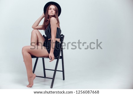 Attractive young model with beautiful make-up and hair-do in stylish black hat and black clothes posing with a chair on a white studio background. Long legs, brown hair, white cyclorama, fashion.