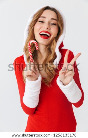 Portrait of young woman 20s wearing Santa Claus red costume smiling and eating Christmas candy isolated over white background