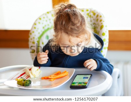 Toddler eats in the high chair while watching movies on the mobile phone. Many children do not want to eat if they do not watch the mobile device or the television.