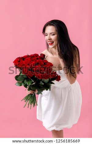 beautiful smiling young woman in a white dress with a large bouquet of red roses on pink background