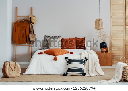 Copy space on white empty wall of trendy bedroom with flower in vase on wooden nightstand, king size bed with autumn colored bedding and wooden ladder