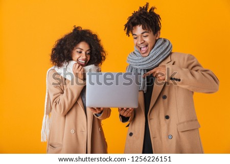 Happy african couple wearing winter clothes standing isolated over yellow background, holding laptop computer, celebrating success