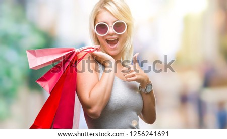 Young beautiful blonde woman shopping holding shopping bags on sales over isolated background happy with big smile doing ok sign, thumb up with fingers, excellent sign