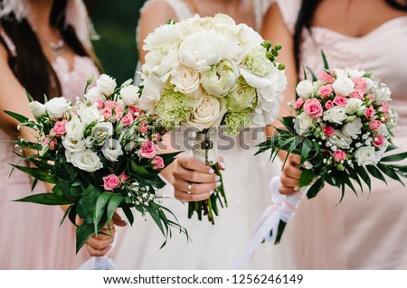 The bride and bridesmaids in an elegant dress is standing and holding hand bouquets of pastel pink flowers and greens with ribbon at nature. Young beautiful girls holds a wedding bouquet outdoors.