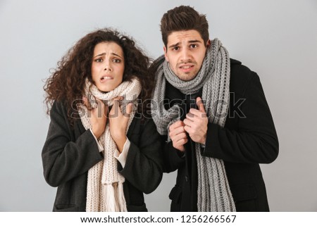 Frozen young couple wearing sweaters and scarfs standing isolated over gray background, shivering