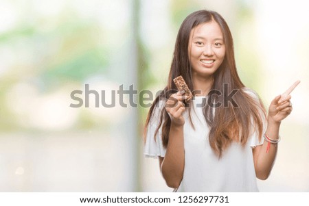 Young asian woman eating chocolate energetic bar over isolated background very happy pointing with hand and finger to the side