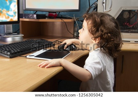 Little 2 3 year old baby girl in white clothers draws at the home computer in graphics drawing tablet. two monitors. The child is holding a mouse.