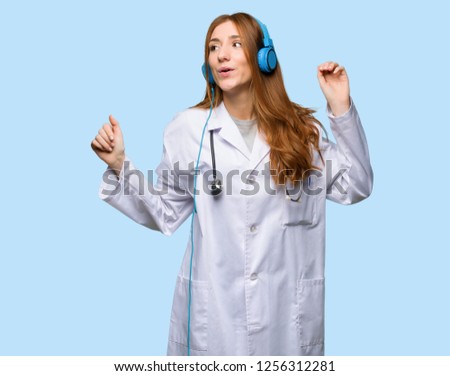 Redhead doctor woman listening to music with headphones and dancing on isolated blue background