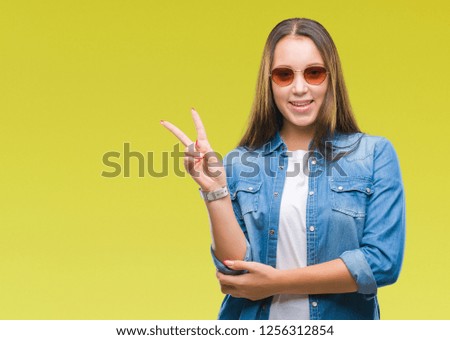 Young beautiful caucasian woman wearing sunglasses over isolated background smiling with happy face winking at the camera doing victory sign. Number two.