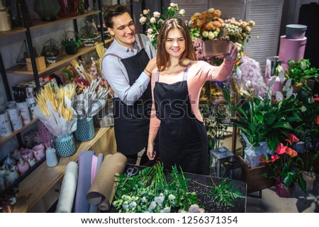 Young femal and male florists stand in room full of flowers and plants. She hold hone. They take selfie. People smile and pose.