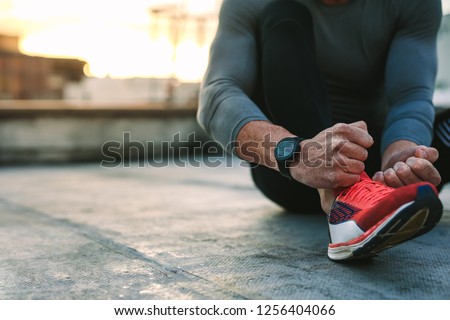 Close up of a fitness man tying lace of his sports shoes. Athlete getting ready for workout wearing shoes sitting on floor. Royalty-Free Stock Photo #1256404066