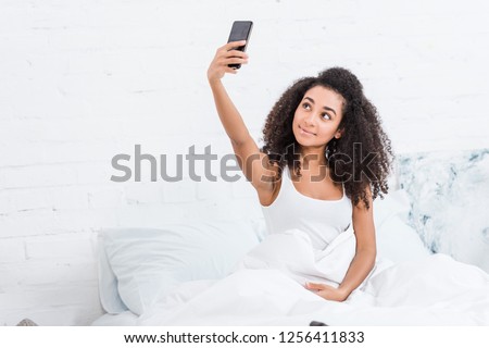 attractive african american girl taking selfie on smartphone in bed during morning time at home