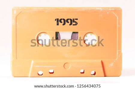 A vintage cassette tape from the 1980s era (obsolete music technology) with the text 1995 printed over it, stencil font. Color: cream, sand. White background.
