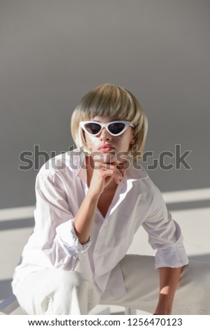 portrait of attractive blonde woman in sunglasses and fashionable white outfit resting chin on hand on white