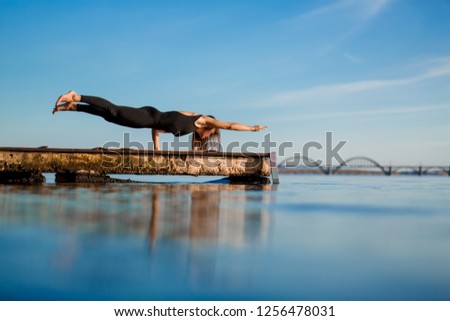 Young woman practicing yoga exercise at quiet wooden pier with city background. Sport and recreation in city rush.