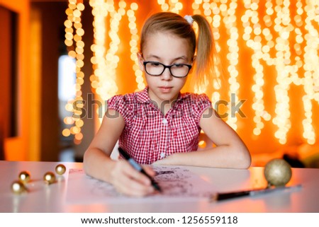 Blonde girl in the pink dress and big black glasses drawing santa claus. Christmas and New Year theme, yellow bokeh.