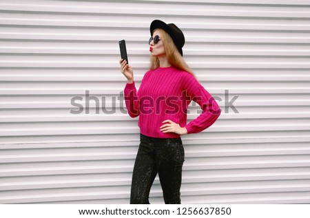 Fashion, technology and people concept - woman taking selfie picture by smartphone blowing red lips sending sweet air kiss in colorful pink knitted sweater, black round hat on white wall background 
