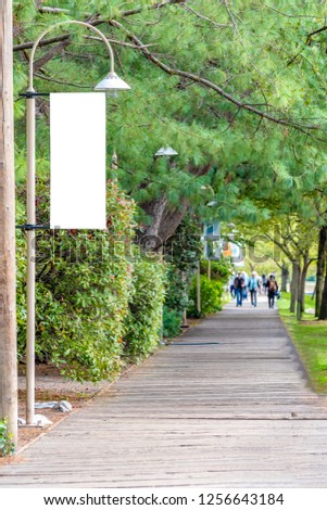 Blank white isolated vertical banner flag mockup signage sign on a streetlight. Public City Park walkway. Unrecognizable people walking in far distance background.