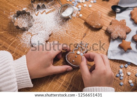 Female hands decorate gingerbread snowman with sugar snowflakes. Spilled powdered sugar and baking molds on the side. Selective focus. Gingerbreads on tablecloth on a wooden table. Christmas season.