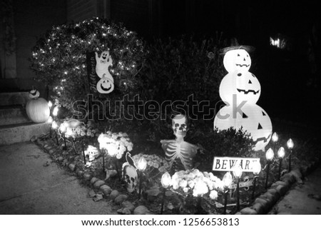 Monochrome Version of Skeleton in Flower Bed with Beware Sign Guarding Front of Townhomes with Plastic Pumpkins Ghost and Other Decorations on Halloween at Night