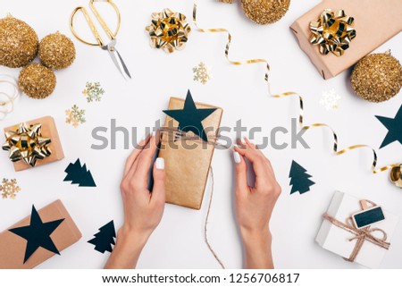 Woman's hands tying gift box with twine and star among festive flat lay frame of Christmas gold and black decorations on white background, view from above.
