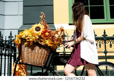 Brunette young girl in a red and white dress holding a classic bicycle full of autumn flowers on a sunny afternoon, spreading happiness and love in the city.