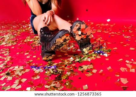 Crop studio shot of young pretty woman wearing boots and evening dress sitting on the floor with confetti and champagne and celebrating New Year, christmas, happy mood, friends