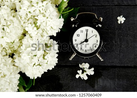 Black alarm clock and white flowers on black background.  Mother or Women Day. Greeting Card. Good Morning breakfast.  Spring. Romantic. Top view. Copy space. Flat lay