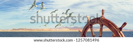 Wooden steering wheel on the sailboat - panorama of sea landscape with mountain seashore and flying birds. Wide view skyline with captains helm on sailling ship and seagulls over for marine concept. Royalty-Free Stock Photo #1257081496